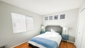 Quarto com cama, mesas de cabeceira, janela com persianas e arte decorativa na parede.