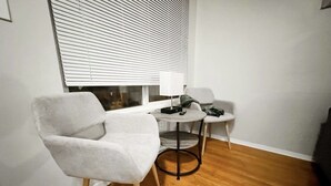 Quarto pequeno e moderno com piso de madeira, uma poltrona branca, uma mesa redonda e uma janela com persianas.
