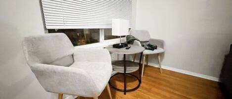 A small, modern room with a wooden floor, a white armchair, a small round table, and a window with blinds.