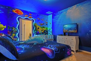 A bedroom with an ocean-themed mural, a bed with a dolphin-shaped pillow, a dresser, and a television on the wall.