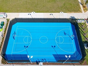 Vista aérea de una cancha de baloncesto azul con dos aros, rodeada por una valla, y contigua a una zona con césped.