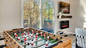 Une pièce avec une table de baby-foot, une cheminée et une grande fenêtre donnant sur les arbres.