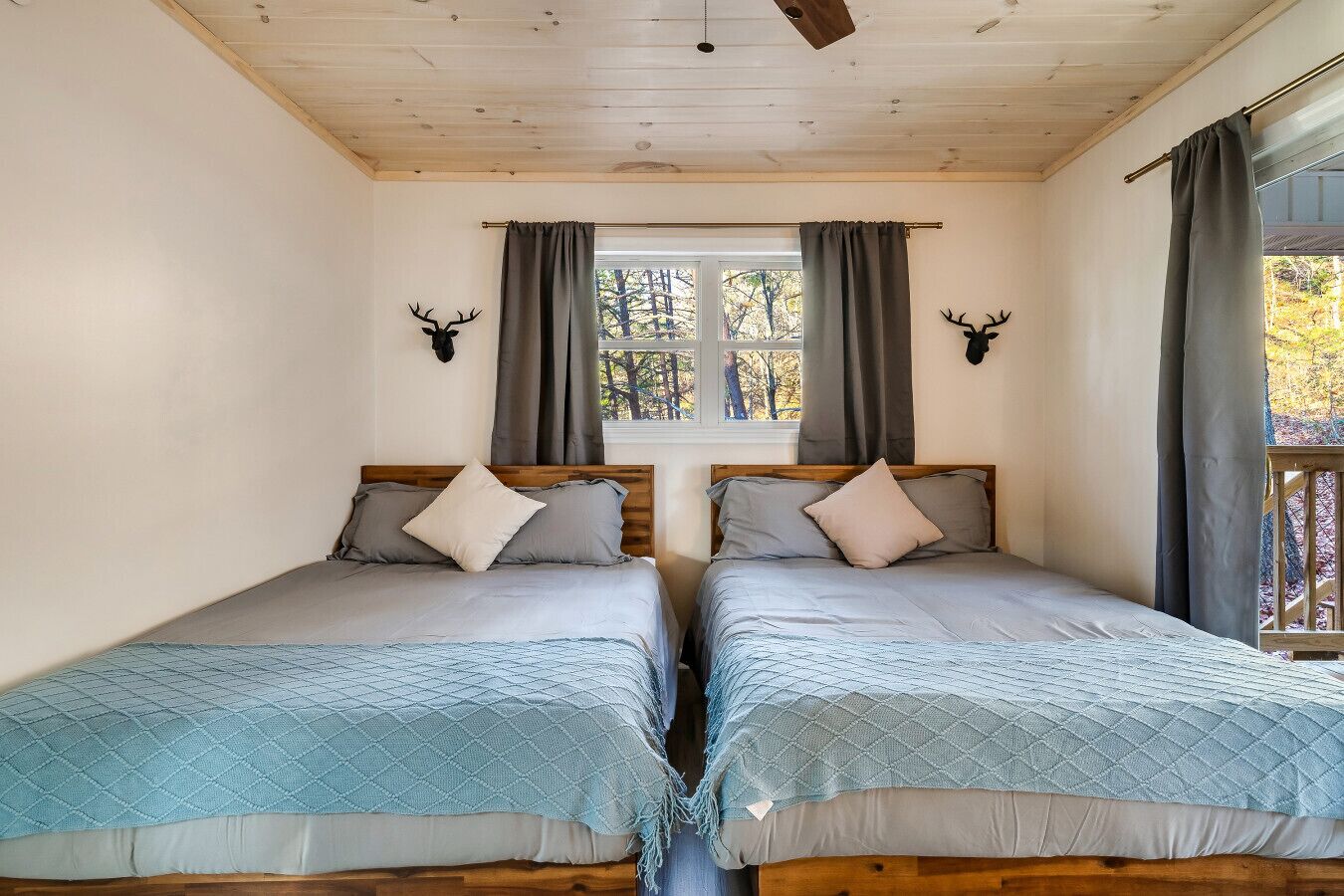 A room with two beds, wooden headboards, a window with curtains, and wall-mounted deer antlers.