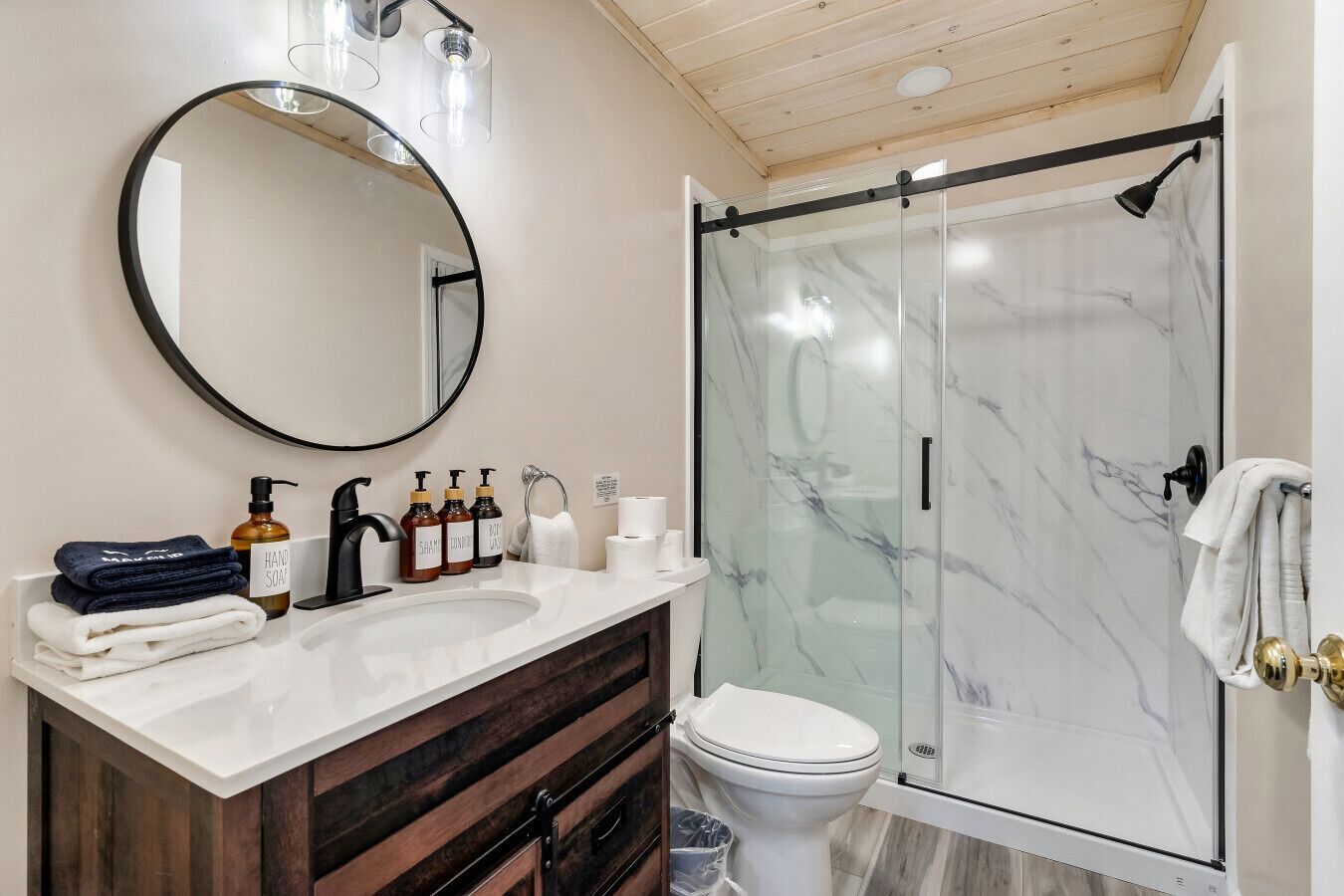 A modern bathroom with a marble shower, a round mirror, and a vanity with toiletries.