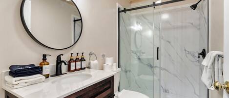 A modern bathroom with a marble shower, a round mirror, and a vanity with toiletries.
