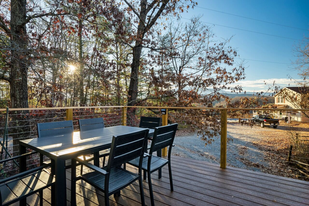 Un terrazzo in legno con tavolo e sedie, affaccia su un paesaggio rurale con alberi e un edificio in lontananza.