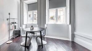 Ett rum med ett runt bord och fyra stolar, stora fönster med gardiner, en hyllsektion i metall och ett mörkt trägolv.