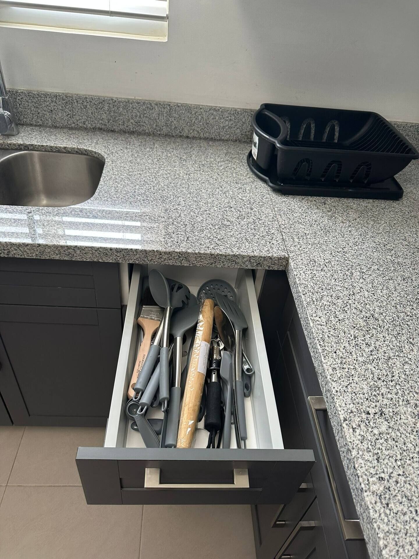 A kitchen with a granite countertop, a stainless steel sink, and a drawer containing kitchen utensils.