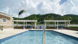Una piscina rectangular amb una superfície d'aigua blava i transparent, envoltada per un solàrium blanc. Hi ha dues pérgoles blanques amb gandules blaves sota. Al fons, hi ha un turó verd i exuberant amb un cel blau i clar.