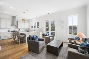 A modern kitchen with white cabinets, a central island, and bar stools. There is a dining area with a gray sofa and a coffee table.