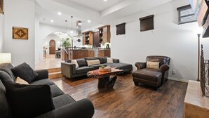 Ein geräumiges Wohnzimmer mit einem großen, dunkelbraunen Ledersofa, einem passenden Sessel und einem Holztisch. Im Hintergrund ist ein Essbereich mit einer Küche und einer Treppe zu einer oberen Ebene zu sehen.