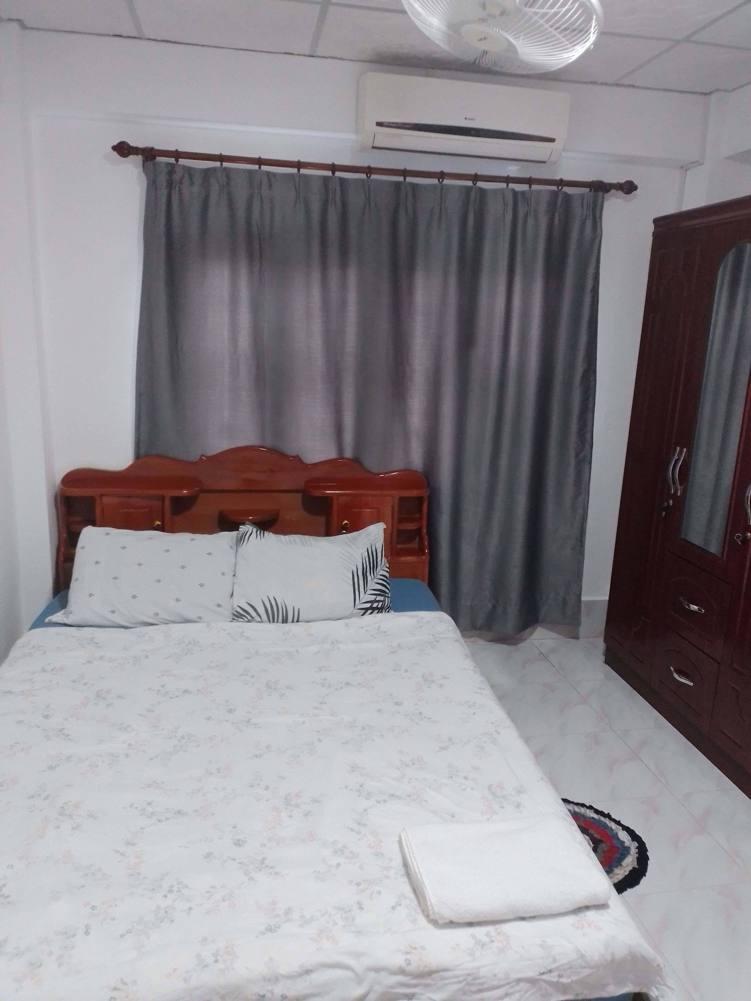 A bedroom with a bed, a wooden headboard, a wardrobe, a ceiling fan, and a window with curtains.