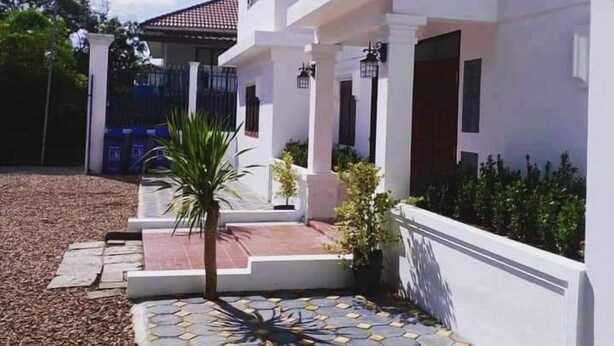 A two-story white building with a tiled entrance area, a small palm tree, and a garden with various plants. - Ban Saylomleng Residence (Vientiane)