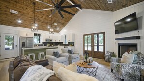 A spacious living room with a high ceiling, a fireplace, and a kitchen area visible through an open door.