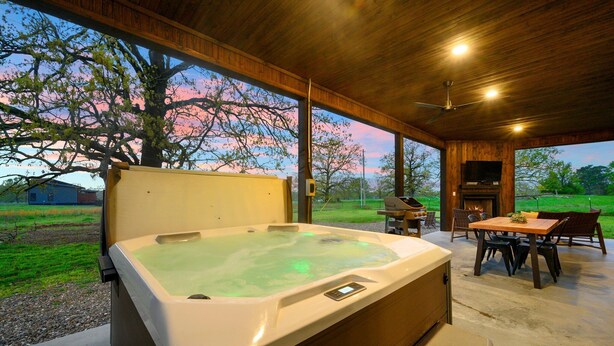A hot tub in a covered outdoor area with a table and chairs, a fireplace, and a view of the surrounding landscape.