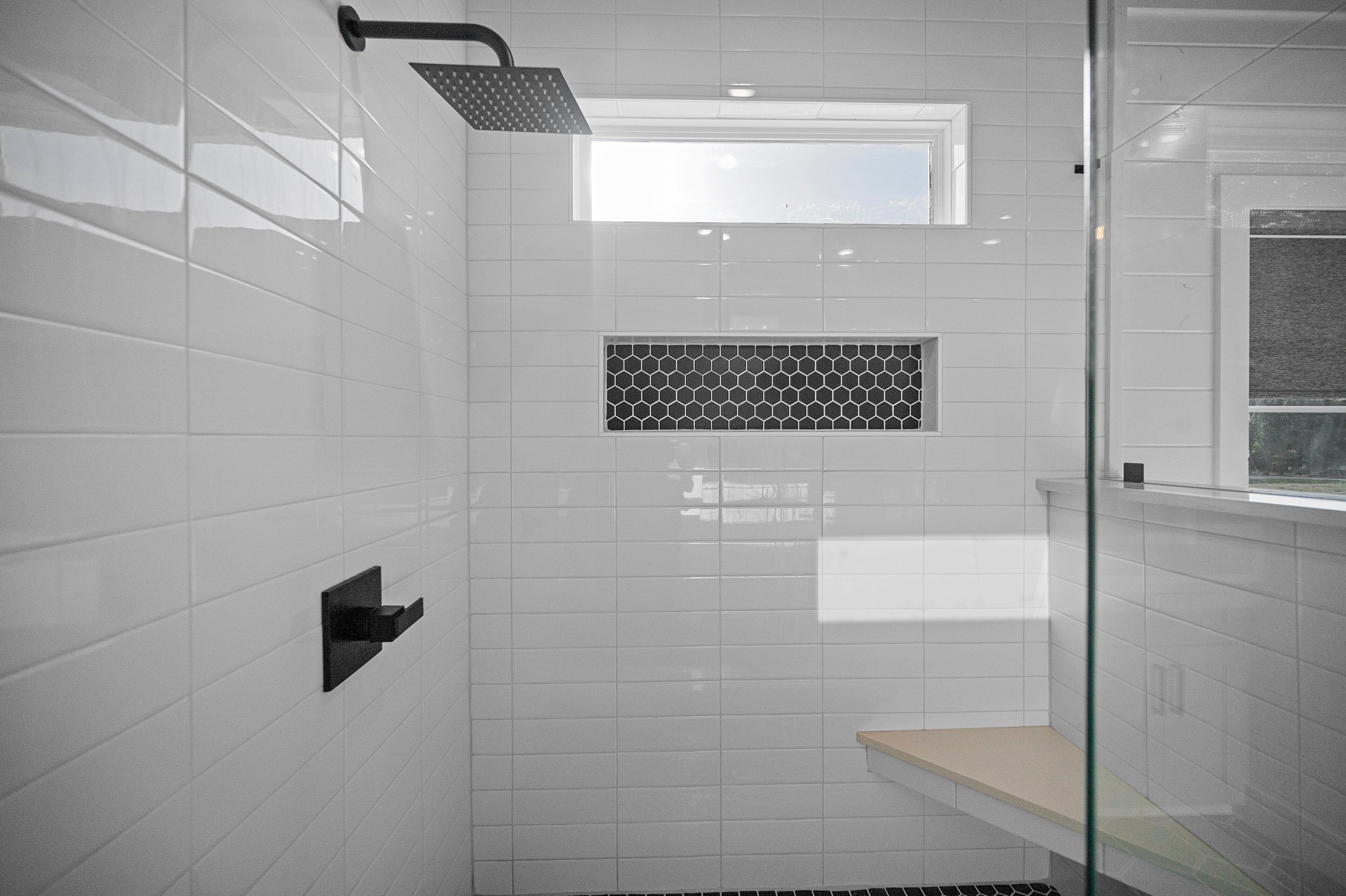 A modern bathroom with white tiles, a showerhead, a small window, and a glass shower enclosure.