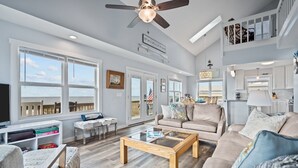 A spacious living room with a large window, a ceiling fan, and a view of the ocean. - Uncle Ed's Watering Hole: 4BR Modern Oceanfront Rental w/ Direct Beach Access & Near Pier (Oak Island)
