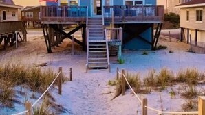 A beachfront house with a balcony, a staircase leading to the entrance, and a sandy beach area. - Uncle Ed's Watering Hole: 4BR Modern Oceanfront Rental w/ Direct Beach Access & Near Pier (Oak Island)