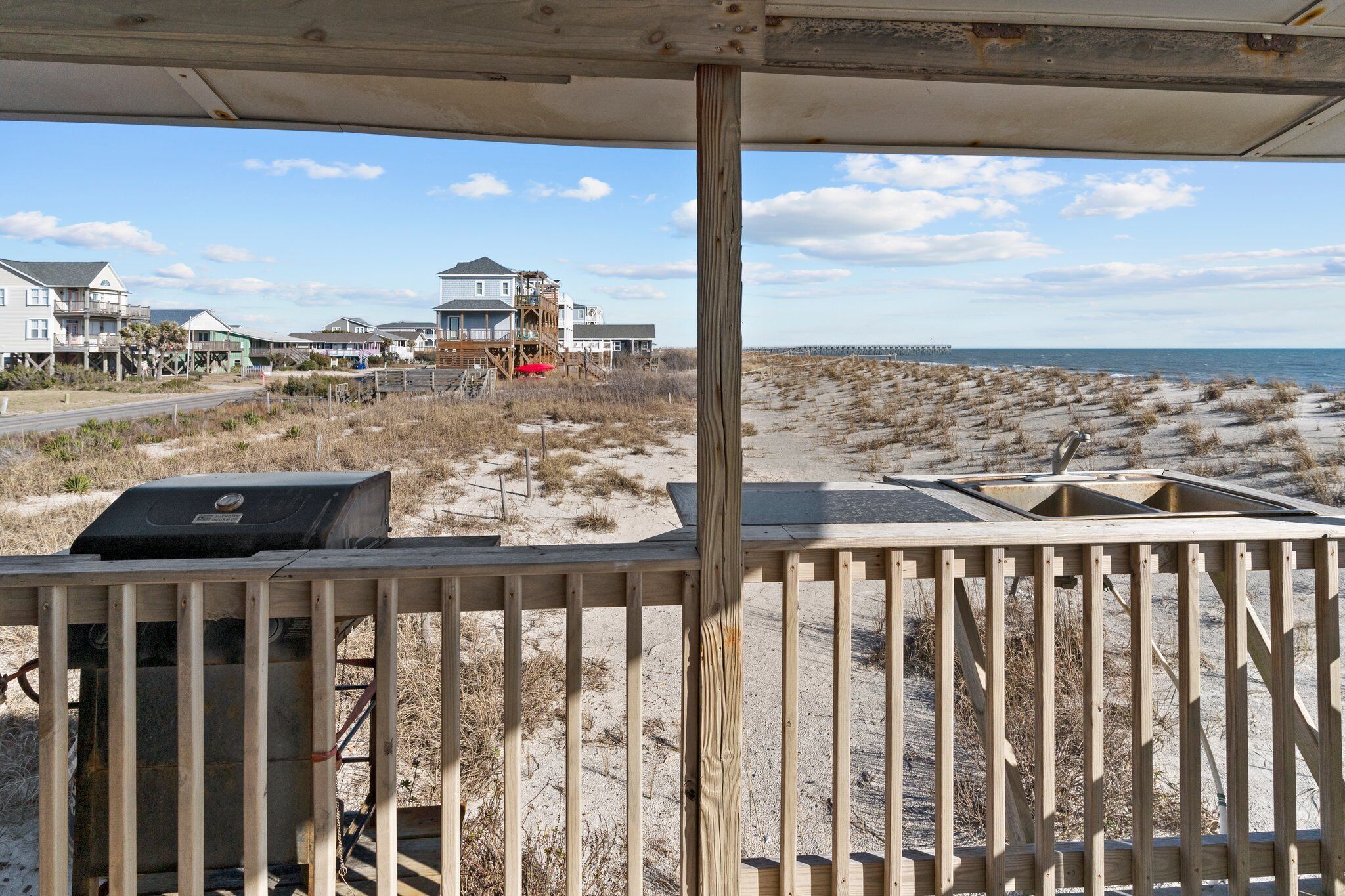 A beach view with a wooden deck, a grill, and a sandy area with sparse vegetation.