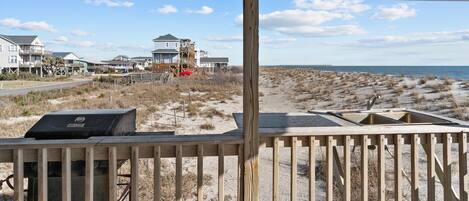 A beach view with a wooden deck, a grill, and a sandy area with sparse vegetation.