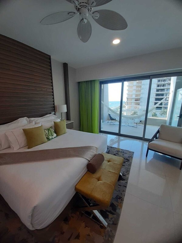 A modern hotel room with a large bed, a sofa, a balcony with a view of the city, and a ceiling fan.