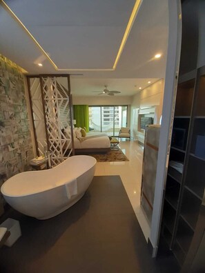 A modern hotel room with a large bathtub, a seating area with a sofa and chairs, a TV, and a balcony with a view.