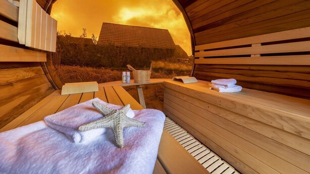 Un sauna avec des bancs en bois, une serviette ornée d’une étoile de mer et un seau rempli de bouteilles, le tout visible à travers une ouverture circulaire.
