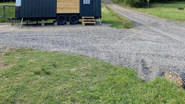 A small, modular cabin with a wooden interior and a black exterior, situated on a gravel path with a grassy area and trees in the background. - The Arkansas Tiny House (Mulberry)