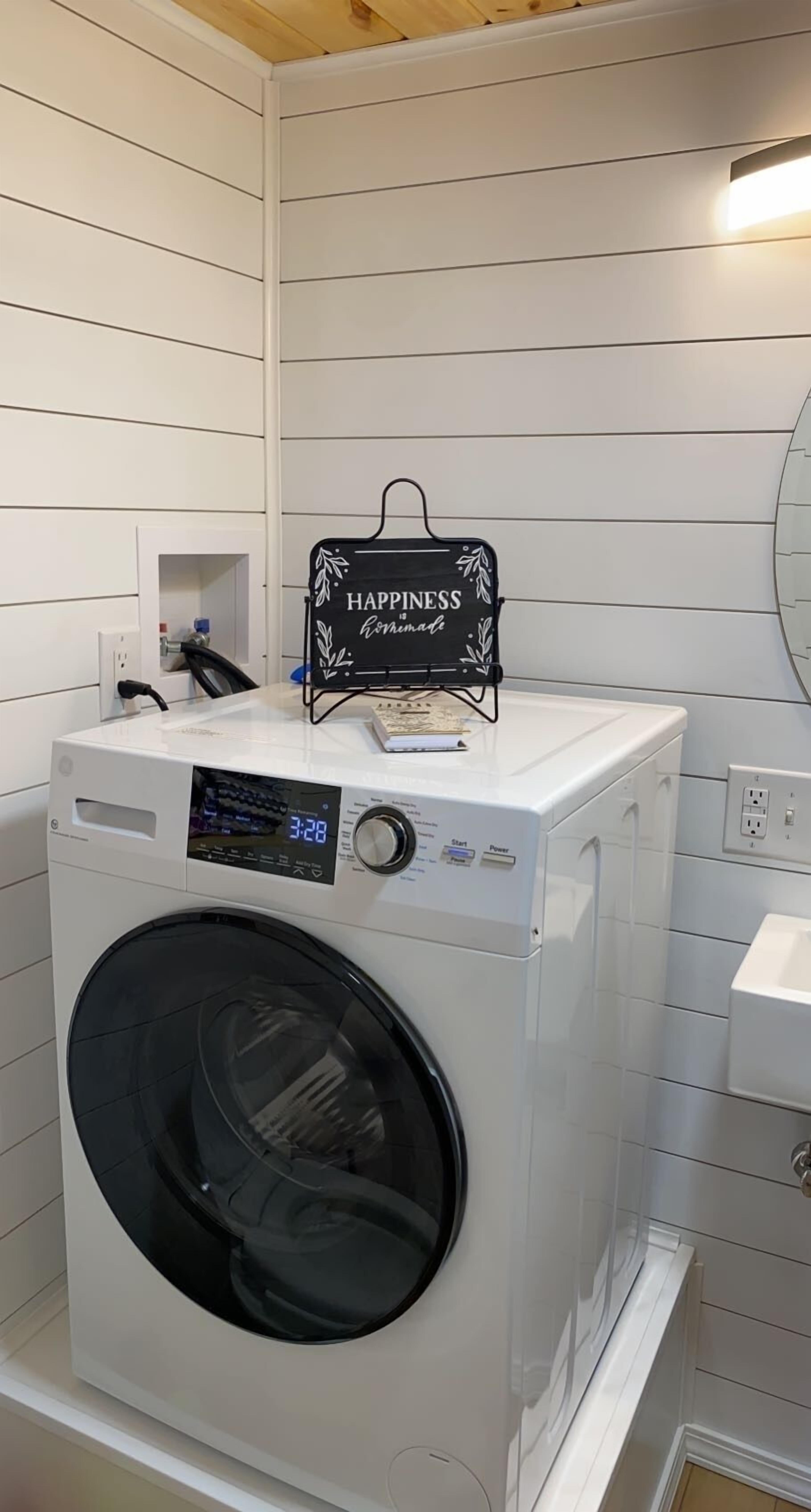 A front-loading washing machine with a digital display, a small shelf with a sign, and a white tiled wall.
