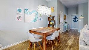A dining area with a wooden table and white chairs, a chandelier, and wall art. - Outdoor Seating, Shared Heated Pool with Water Slide, Golf Cart Access (Port Aransas)