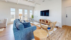 A spacious living room with a blue sofa, wicker chairs, a flat-screen TV, and a wooden coffee table. - Private Heated Pool, Golf Cart Included, Pet Friendly (Port Aransas)