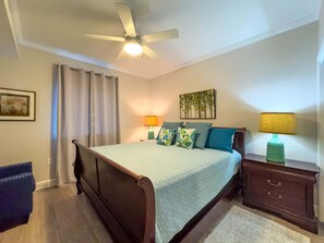 A bedroom with a bed, a nightstand, a lamp, a ceiling fan, and a painting on the wall.
