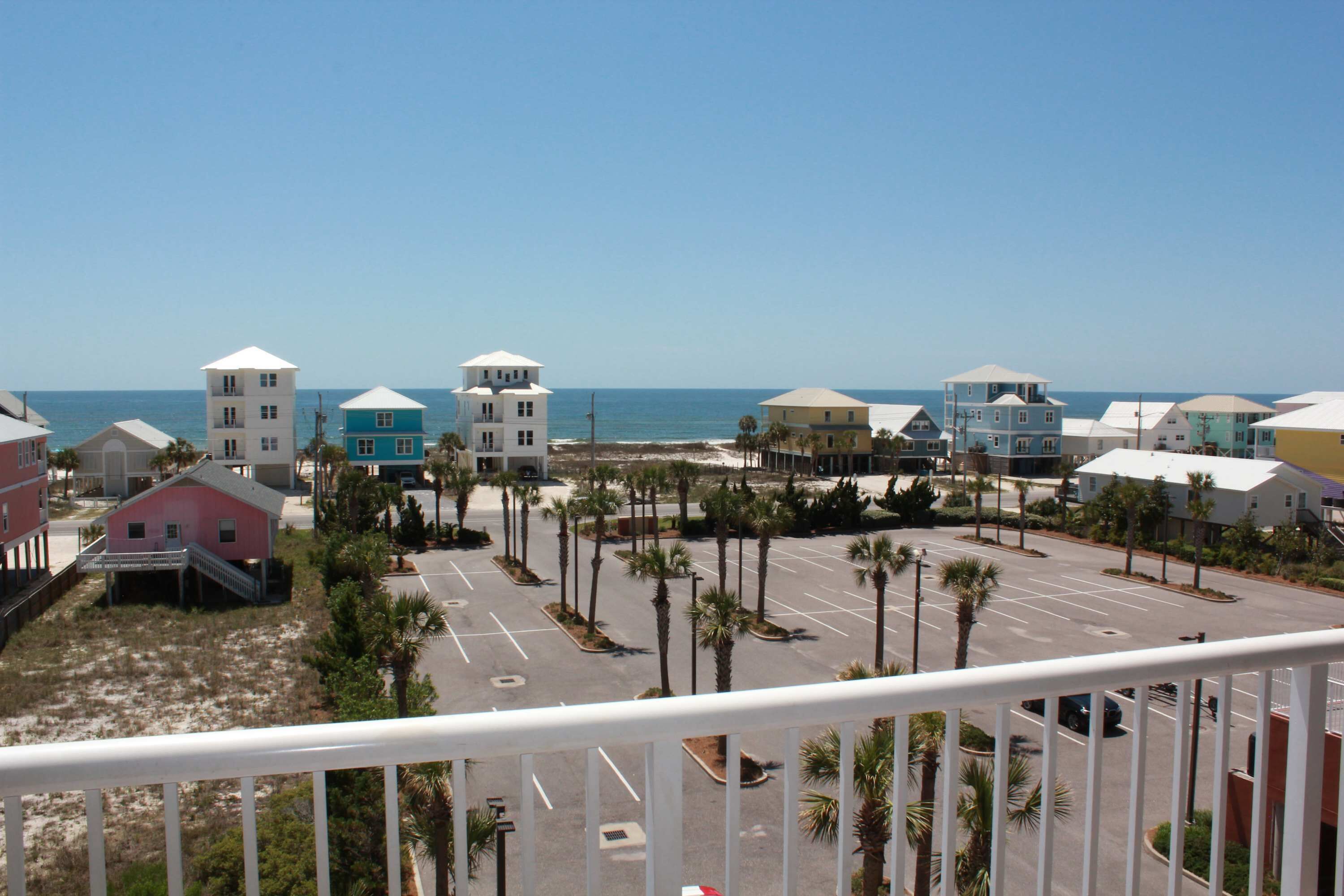A coastal view of a parking lot with several buildings, including a pink house and a blue building with a white roof, near the beach.