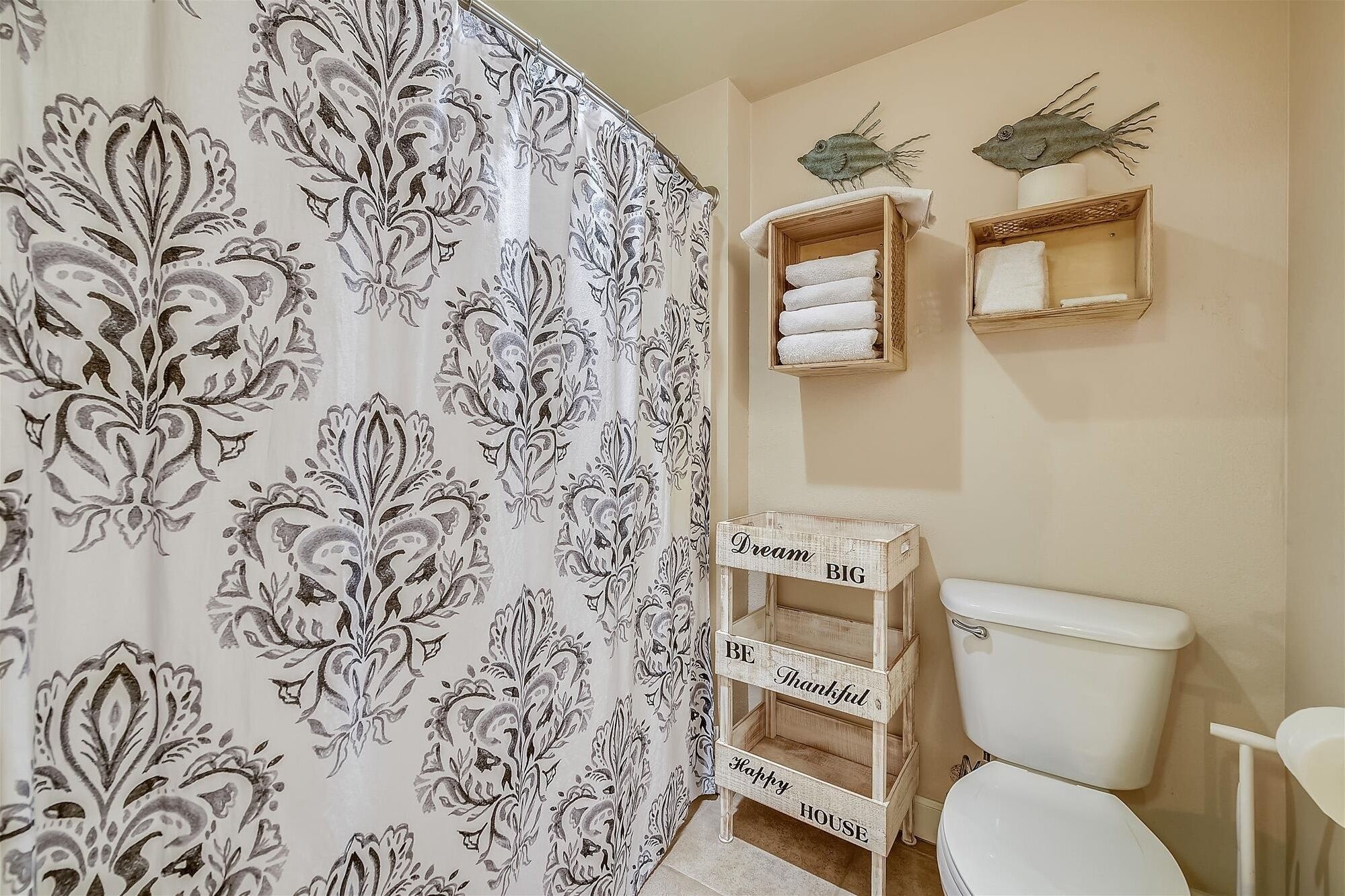 A bathroom with a toilet, a shower curtain with a floral pattern, and wooden shelves with towels and decorative fish.