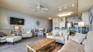 Un salon comprenant un canapé, des fauteuils, une table basse, un téléviseur, un ventilateur de plafond, et un coin cuisine à l’arrière-plan.