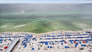 Une plage bordée de rangées de parasols bleus et de chaises de plage, une longue structure en bois, et l’océan en arrière-plan.