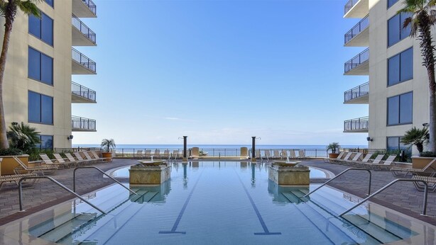 Une piscine à l’eau d’un bleu limpide, entourée de chaises longues et de palmiers, avec vue sur une plage.