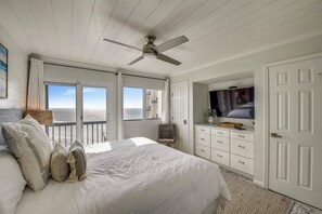 Relax in this serene bedroom with a large bed, wicker chair, and flat-screen TV, offering a stunning ocean view.