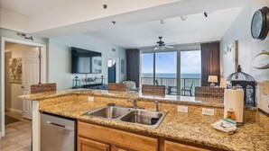 A kitchen with granite countertops, a stainless steel sink, and wooden cabinets. There is a large window offering a view of the ocean. - Shores of Panama 1418 - 2 Bedroom+Bunkroom ! Updated with a View! Free Fun! 2 Condo by RedAwning (Panama City Beach)