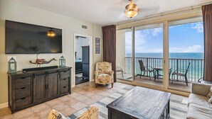 A spacious living room with a large TV, a wooden coffee table, and a view of the ocean through a balcony. - Shores of Panama 1418 - 2 Bedroom+Bunkroom ! Updated with a View! Free Fun! 2 Condo by RedAwning (Panama City Beach)