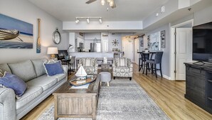 A spacious living room with a gray sofa, patterned armchairs, a coffee table, and a dining area with bar stools. A kitchen is visible in the background, and a ceiling fan is mounted above the seating area. - Shores of Panama 1011 - 1 Bedroom+Bunks . Free Fun! 1 Condo by RedAwning (Panama City Beach)