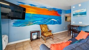 A living room with a large wall mural of a sunset over the ocean, a television mounted on the wall, a dining area with a table and chairs, and a sofa with colorful cushions. - Shores Of Panama 1506, Perfect For A Great Couples Getaway! Free Fun! Reserved Parking 1 Bedroom Condo by RedAwning (Panama City Beach)