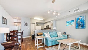 A living room with a blue-striped sofa, a wooden coffee table, and a dining area with a table and chairs. - Shores Of Panama 726 - 1bd+bunks, Sleeps 6. Sunset View! Reserved Parking Space! Free Fun! 1 Bedroom Condo by RedAwning (Panama City Beach)