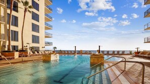 A pool area with clear blue water, surrounded by lounge chairs and palm trees, with a view of the ocean and a building with balconies. - Origin at Seahaven 1036/1038 - 2 Bedroom Unit 2 Home by RedAwning (Panama City Beach)