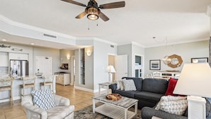 A spacious living room with a dark grey sofa, a wooden coffee table, and a patterned armchair. A ceiling fan and a dining area with a white table and chairs are visible. - Gulf Crest 2005 - 2 Bedroom W/Fantastic View! Family Friendly! Sleeps 8 Condo by RedAwning (Panama City Beach)