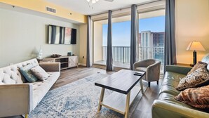 Een moderne woonkamer met een bank, een salontafel en een televisie die aan de muur is bevestigd. Er is een balkon met uitzicht op de stad.