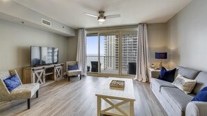 A modern living room with a TV, seating, a coffee table, and a balcony with a view of buildings. - Shores Of Panama 1426 1 Bd+bunk Room, Sleeps 6. Renovated! Sunset View! Free Fun! 1 Bedroom Condo by RedAwning (Panama City Beach)