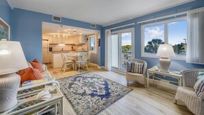 A spacious living room with a large window, a kitchen with white cabinets, and a dining area with wicker furniture. - Recently Renovated 2bd, Sleeps 6 Pinnacle Port, W/ Boat Ramp 2 Bedroom Condo by RedAwning (Panama City Beach)