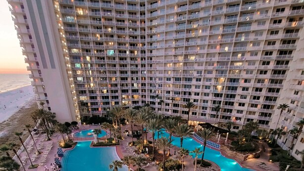 Um hotel grande com vários andares, uma piscina central e palmeiras ao redor.