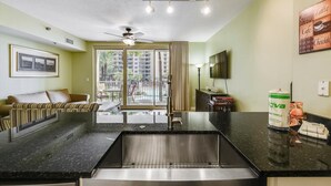A modern kitchen with a stainless steel sink, a dining area with a sofa and chairs, a ceiling fan, and a view of a building through a large window. - Shores of Panama 204 - 2nd Floor 2 Bedroom . Same Floor Reserved Parking. Free Fun 2 Condo by RedAwning (Panama City Beach)